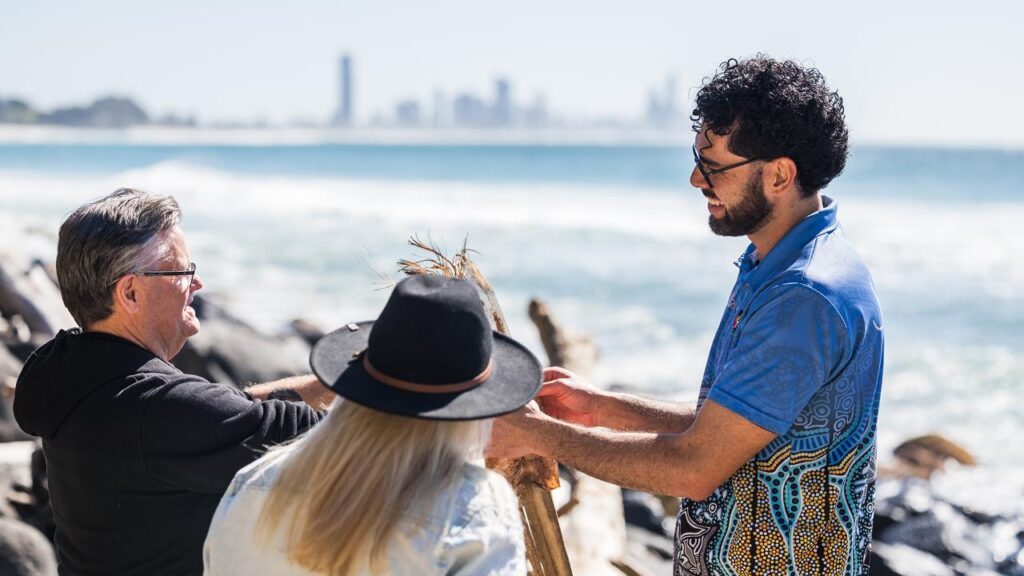 Three people stand on rocky coastline near the ocean, talking and smiling. One holds a stick. City skyline is visible in the background under a clear sky.