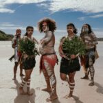 Five people in traditional clothing and body paint stand on a sandy beach holding green foliage, with blue sky and greenery in the background.