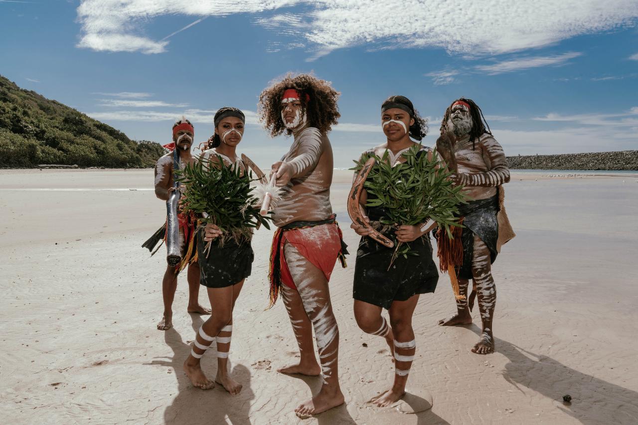 Five people in traditional clothing and body paint stand on a sandy beach holding green foliage, with blue sky and greenery in the background.