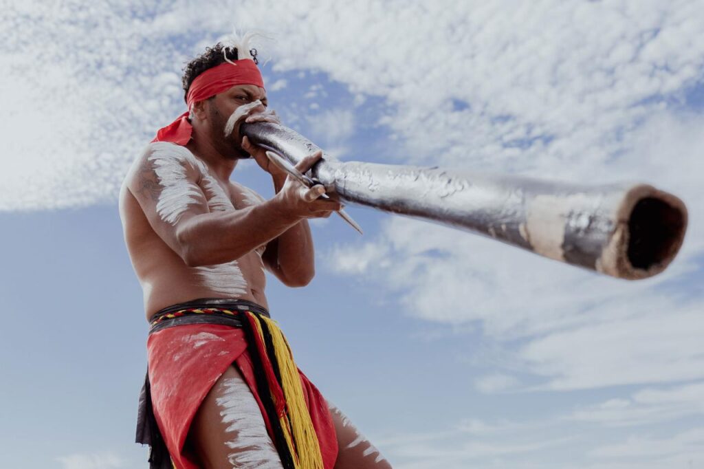 A person wearing traditional attire and body paint plays a didgeridoo outdoors under a partly cloudy sky.
