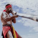 A person wearing traditional attire and body paint plays a didgeridoo outdoors under a partly cloudy sky.