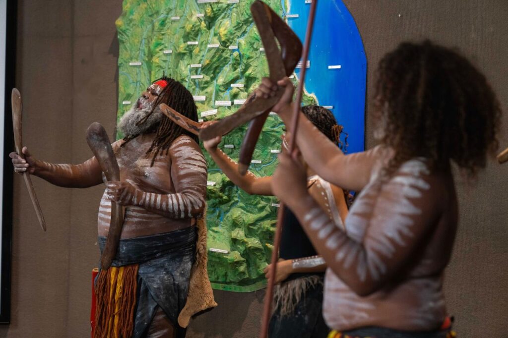 A group of people with body paint and traditional attire perform a cultural dance with boomerangs in front of a green backdrop.