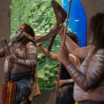 A group of people with body paint and traditional attire perform a cultural dance with boomerangs in front of a green backdrop.