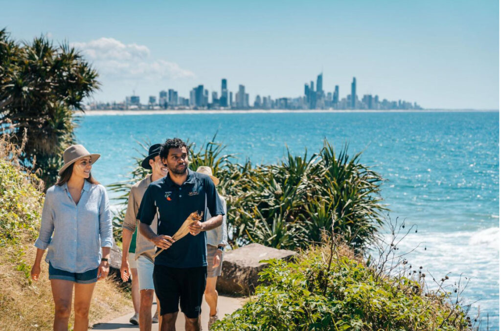 Three people walk along a coastal path with greenery, with the ocean on one side and a distant city skyline under a clear blue sky.