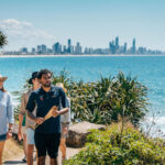 Three people walk along a coastal path with greenery, with the ocean on one side and a distant city skyline under a clear blue sky.
