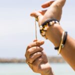 Close-up of hands holding a string, with one hand wearing colorful beaded bracelets, against a blurred outdoor background with water and sky.