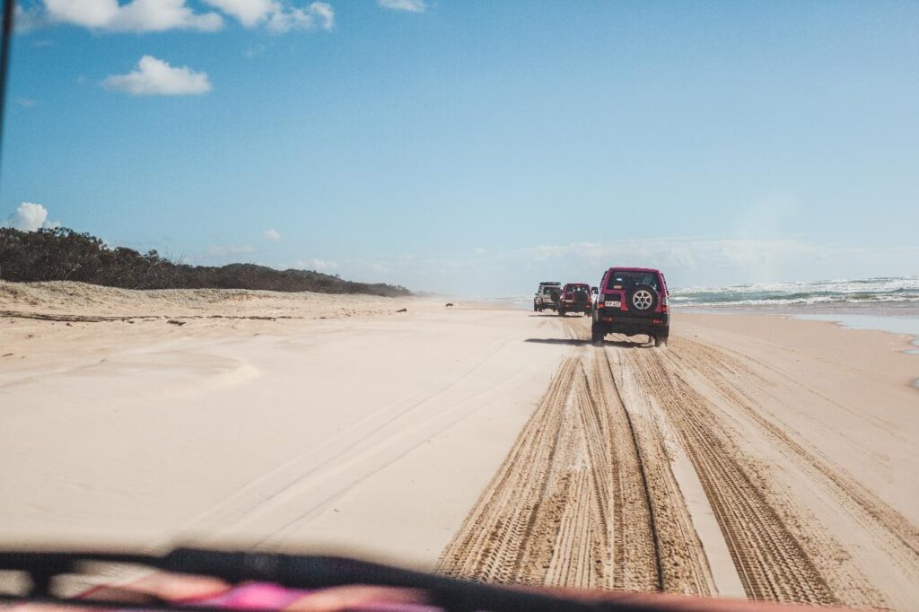 Three SUVs drive in a line on a sandy beach, leaving tire tracks. The sky is mostly clear with a few clouds, and the ocean is visible on the right side.