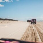 Three SUVs drive in a line on a sandy beach, leaving tire tracks. The sky is mostly clear with a few clouds, and the ocean is visible on the right side.