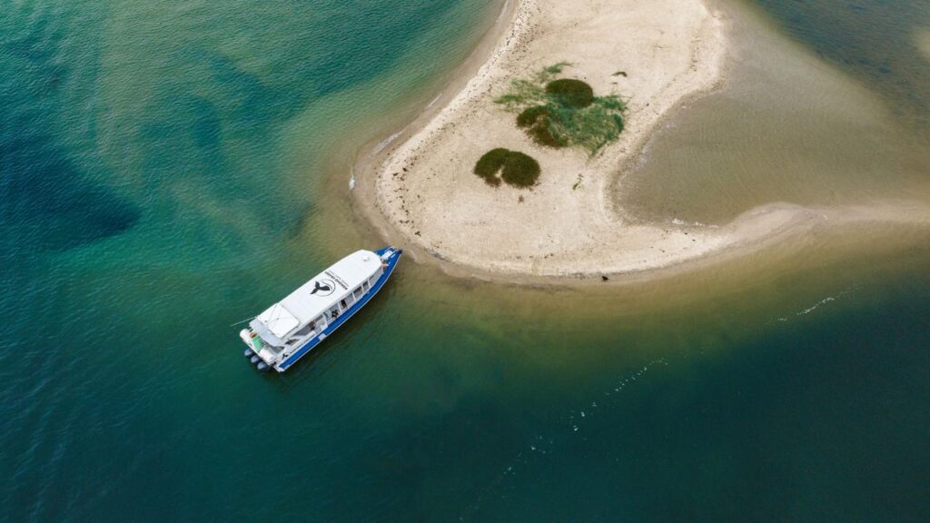 A blue and white boat is docked beside a small sandy island with sparse vegetation, surrounded by clear blue-green water.
