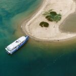 A blue and white boat is docked beside a small sandy island with sparse vegetation, surrounded by clear blue-green water.