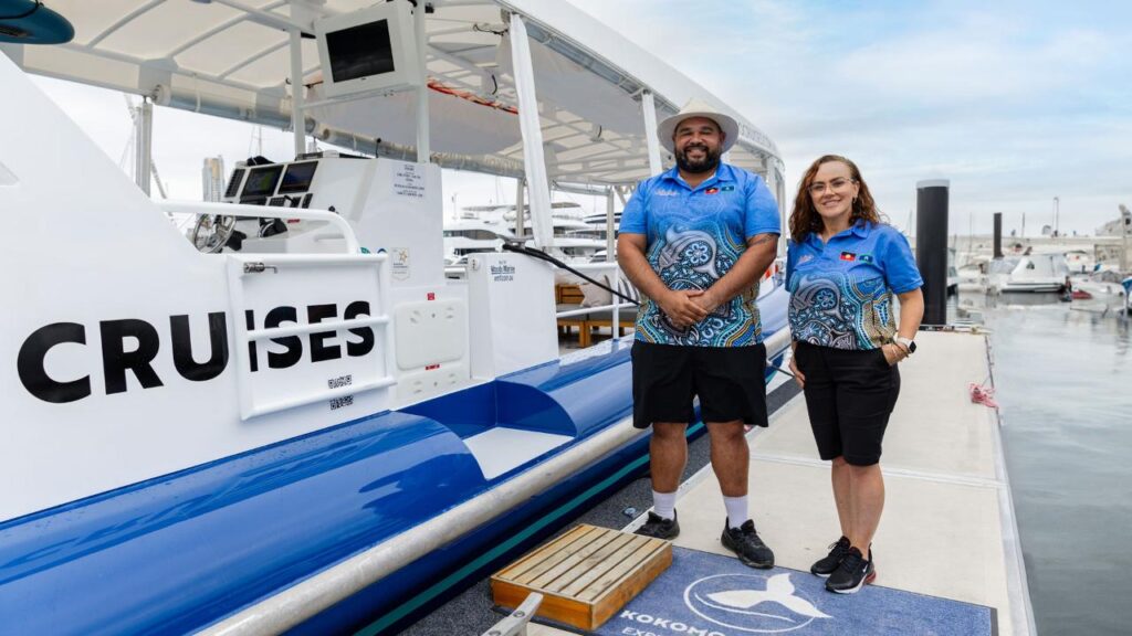 Two people in matching blue uniforms stand on a dock next to a boat labeled "CRUISES," with marina and boats visible in the background.