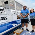 Two people in matching blue uniforms stand on a dock next to a boat labeled "CRUISES," with marina and boats visible in the background.