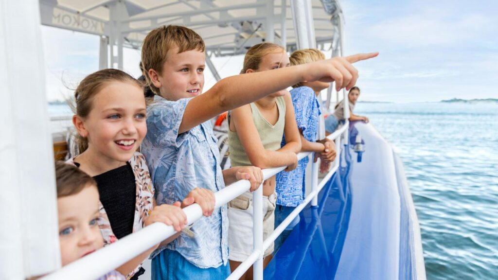 A group of children and adults stand at the railing of a boat, looking and pointing out over the water on a clear day.