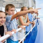 A group of children and adults stand at the railing of a boat, looking and pointing out over the water on a clear day.