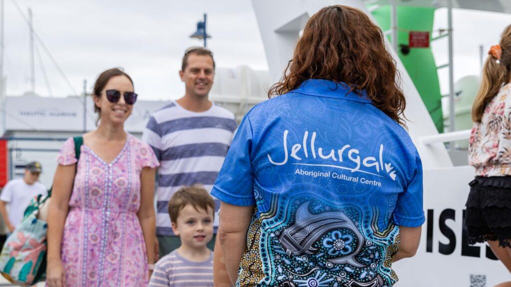 A person wearing a "Jellurgal Aboriginal Cultural Centre" shirt faces a smiling family of three at an outdoor event near boats.
