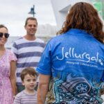 A person wearing a "Jellurgal Aboriginal Cultural Centre" shirt faces a smiling family of three at an outdoor event near boats.