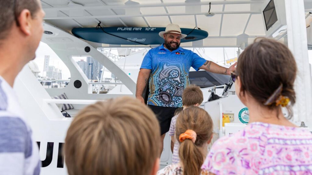 A man in a blue shirt and hat stands on a boat, speaking to a small group of people, including children, with a city skyline in the background.