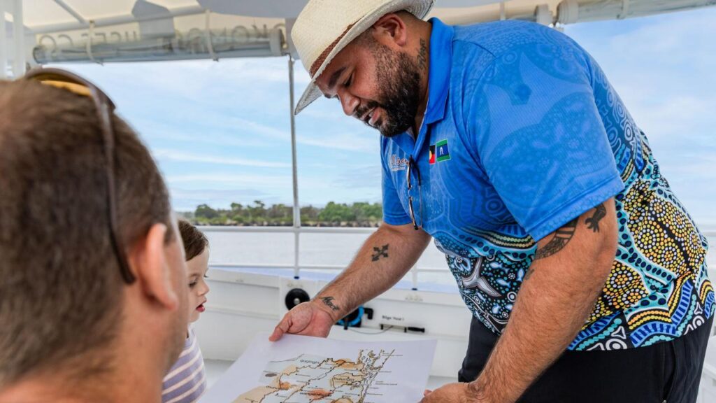 A man in a blue patterned shirt and white hat shows a map to two people on a boat, with water and trees visible in the background.