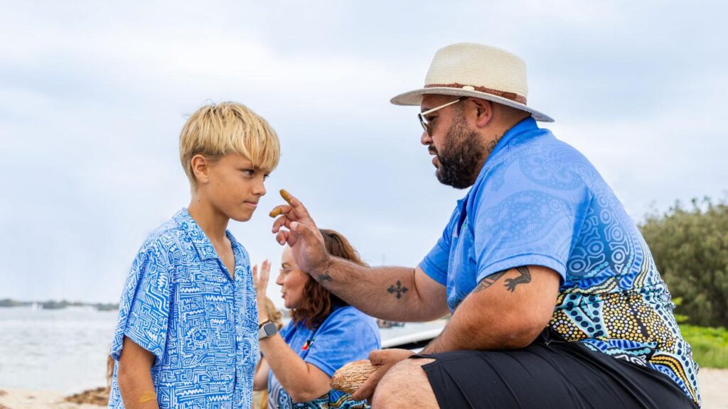 An adult applies ochre to a boy’s forehead at the beach while another person sits in the background. All wear blue patterned clothes.