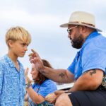 An adult applies ochre to a boy’s forehead at the beach while another person sits in the background. All wear blue patterned clothes.