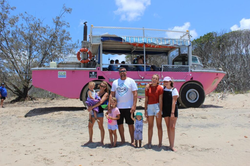 A group of six people, including four children and two adults, stand on sandy ground in front of a pink amphibious tour vehicle with trees in the background.
