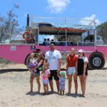 A group of six people, including four children and two adults, stand on sandy ground in front of a pink amphibious tour vehicle with trees in the background.