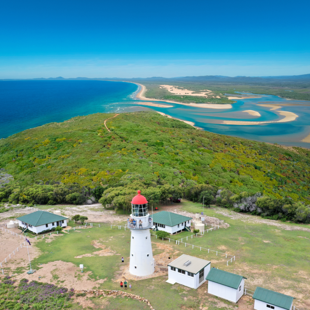A white lighthouse with a red roof is surrounded by small buildings and greenery, overlooking a coastline with sandy beaches and blue water under a clear sky.