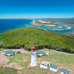 A white lighthouse with a red roof is surrounded by small buildings and greenery, overlooking a coastline with sandy beaches and blue water under a clear sky.