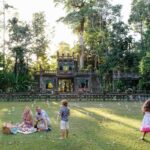 A family has a picnic on the grass in front of a historic stone structure surrounded by trees, while two children play nearby and bubbles float in the air.