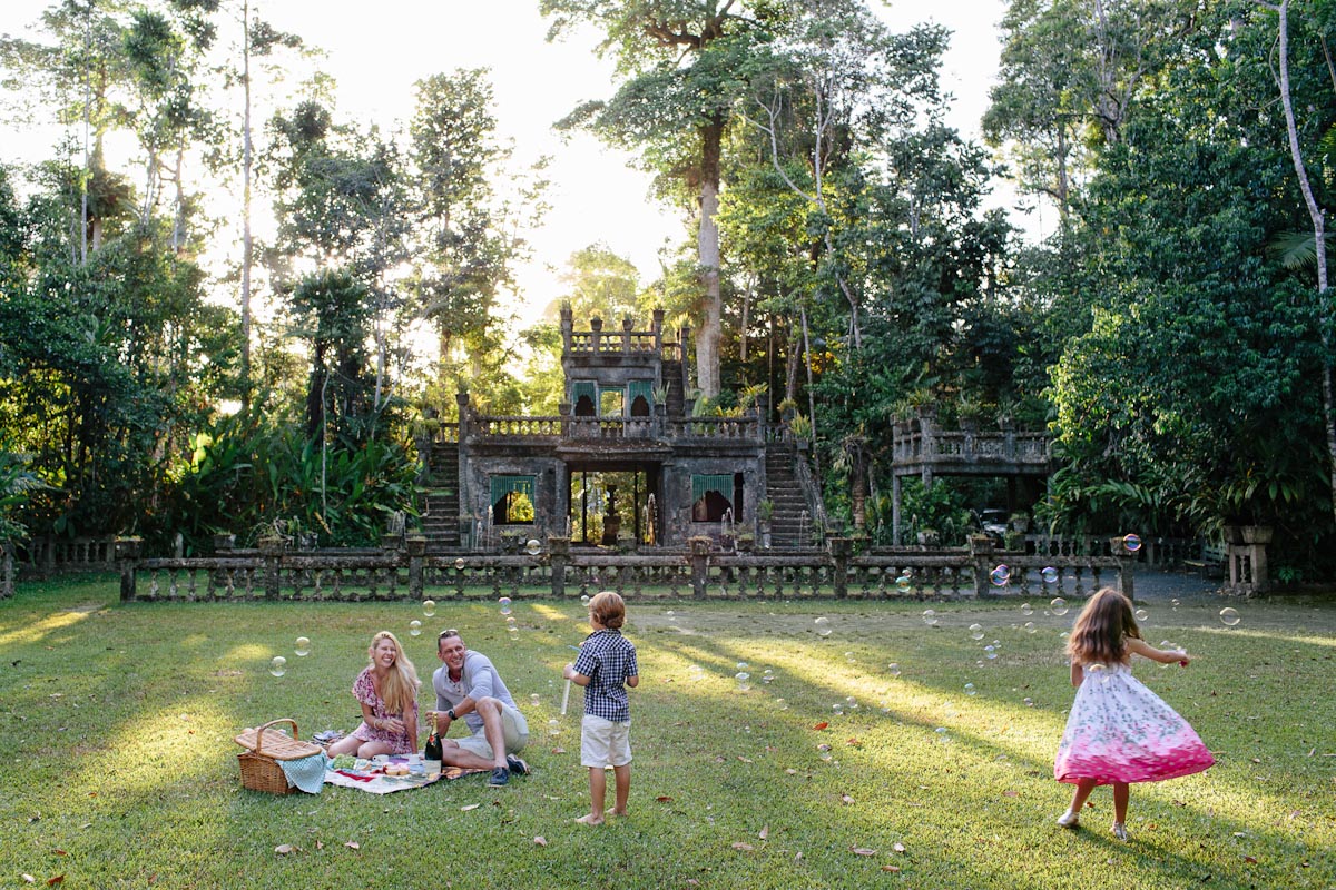 A family has a picnic on the grass in front of a historic stone structure surrounded by trees, while two children play nearby and bubbles float in the air.