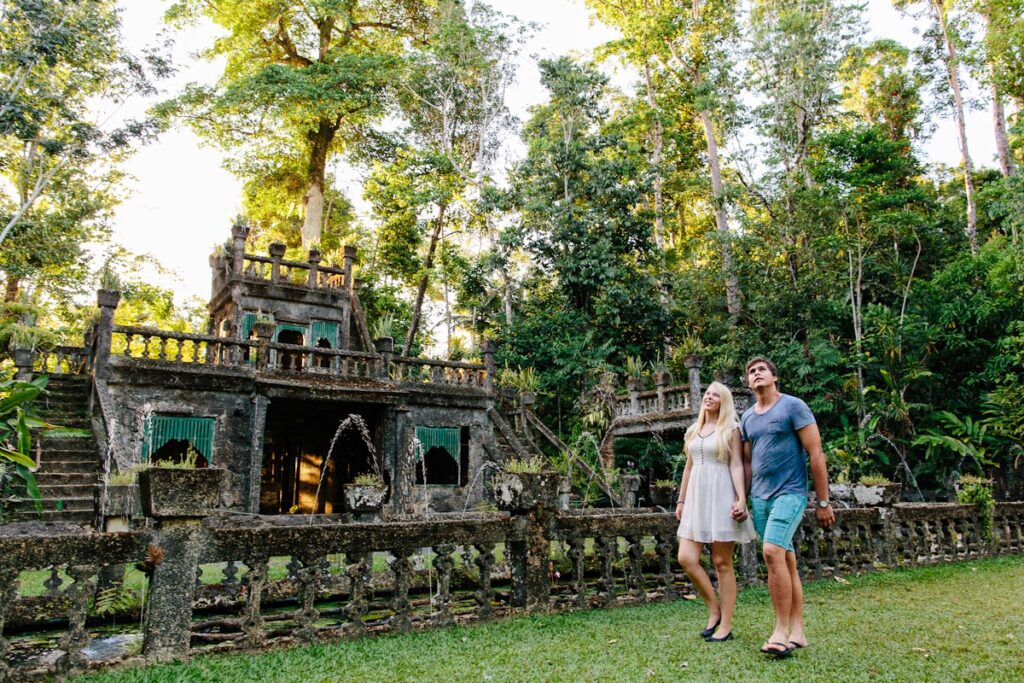 A couple walks hand in hand on a grassy area in front of a historic stone structure surrounded by lush greenery and trees.