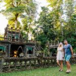 A couple walks hand in hand on a grassy area in front of a historic stone structure surrounded by lush greenery and trees.