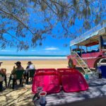 A group of people sit on plastic chairs near a pink beach vehicle under tree shade, with trays and cups on a table and the ocean in the background.