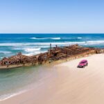 A rusty shipwreck sits partly submerged in shallow water near the shore of a sandy beach, with a pink SUV parked nearby.