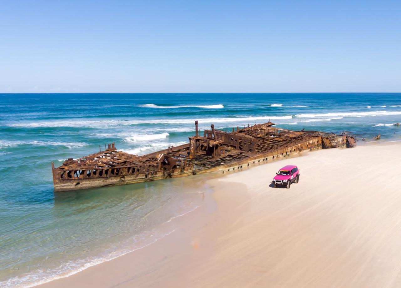 A rusty shipwreck sits partly submerged in shallow water near the shore of a sandy beach, with a pink SUV parked nearby.