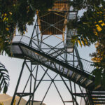 Tall metal observation tower with multiple staircases and platforms, surrounded by trees, overlooking distant hills under a clear sky.