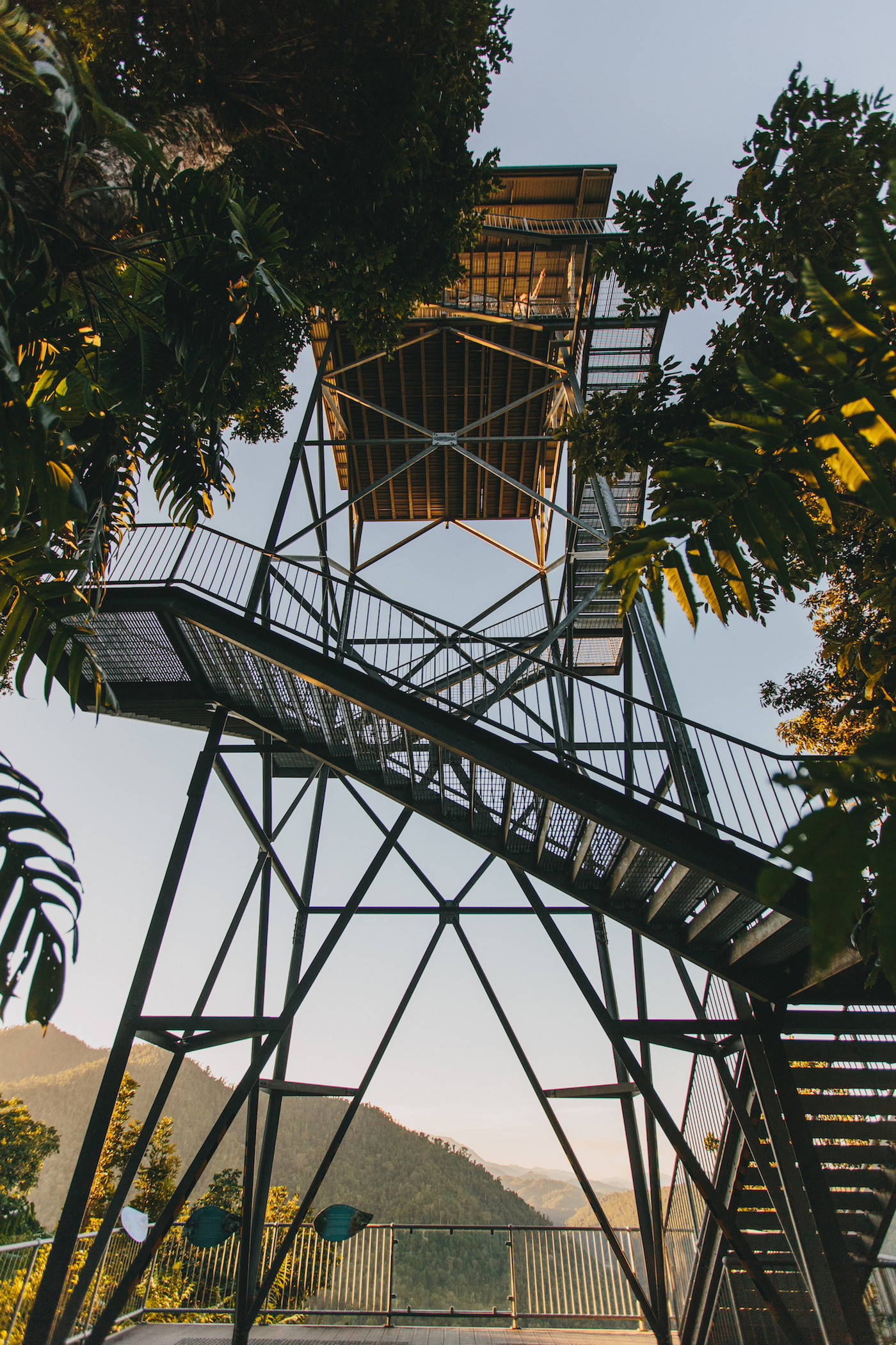 Tall metal observation tower with multiple staircases and platforms, surrounded by trees, overlooking distant hills under a clear sky.
