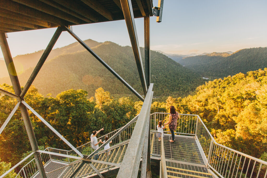 Three people stand on a metal observation platform overlooking a forested mountain landscape during sunset.