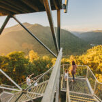 Three people stand on a metal observation platform overlooking a forested mountain landscape during sunset.