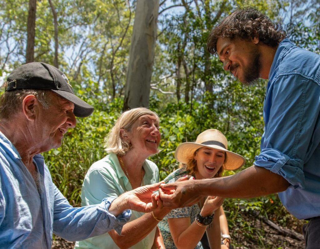 Four adults outdoors examine a small object in one person's hand, smiling and interacting in a forested area on a sunny day.