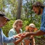 Four adults outdoors examine a small object in one person's hand, smiling and interacting in a forested area on a sunny day.