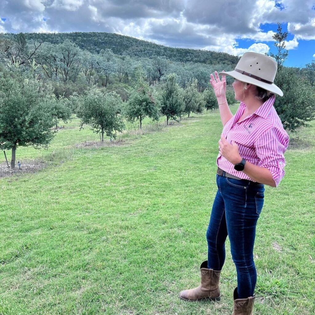 A woman in a pink shirt, jeans, and boots stands on grass, facing and gesturing toward a tree-covered hill under a partly cloudy sky.