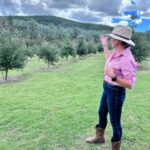 A woman in a pink shirt, jeans, and boots stands on grass, facing and gesturing toward a tree-covered hill under a partly cloudy sky.