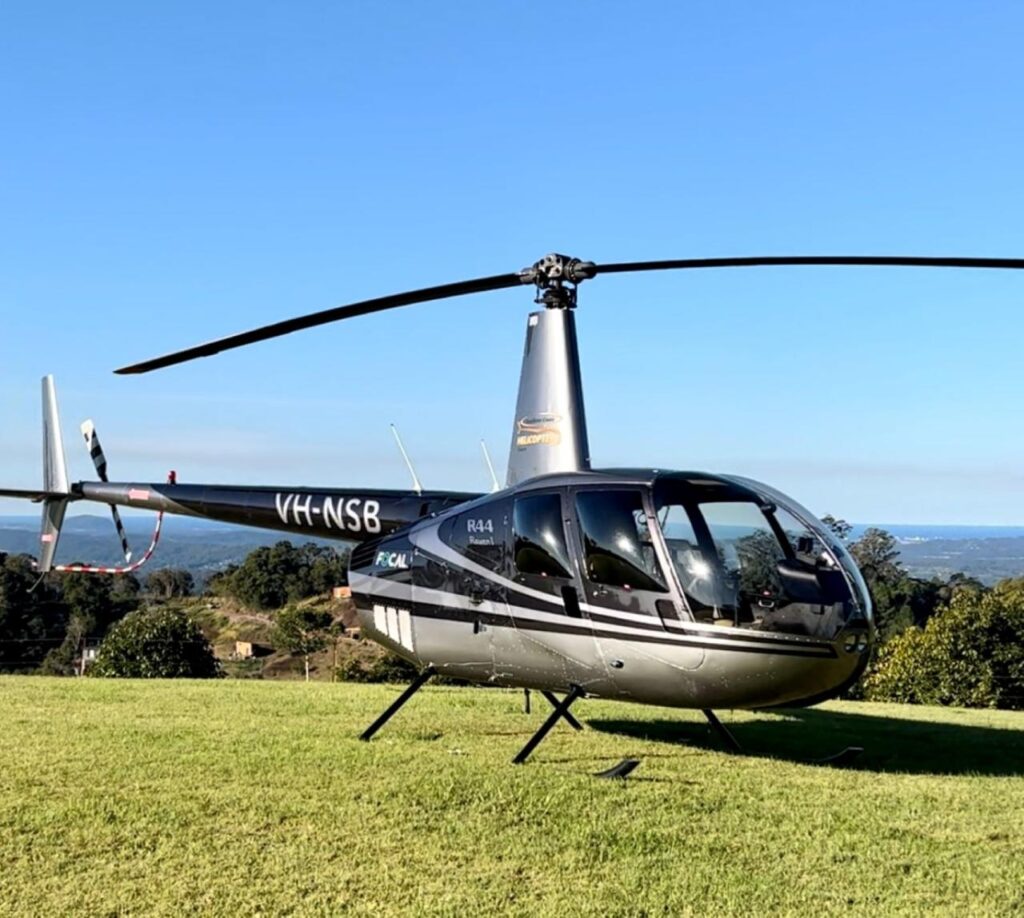 A gray and black Robinson R44 helicopter with registration VH-NSB is parked on a grassy field with trees and distant hills in the background under a clear blue sky.