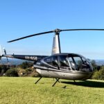 A gray and black Robinson R44 helicopter with registration VH-NSB is parked on a grassy field with trees and distant hills in the background under a clear blue sky.