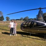 A man and woman stand side by side on grass next to a black helicopter, with clear blue sky and a vineyard in the background.