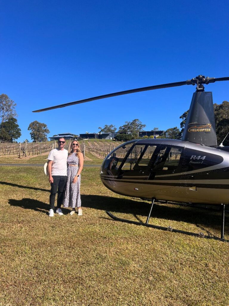 A man and woman stand side by side on grass next to a black helicopter, with clear blue sky and a vineyard in the background.