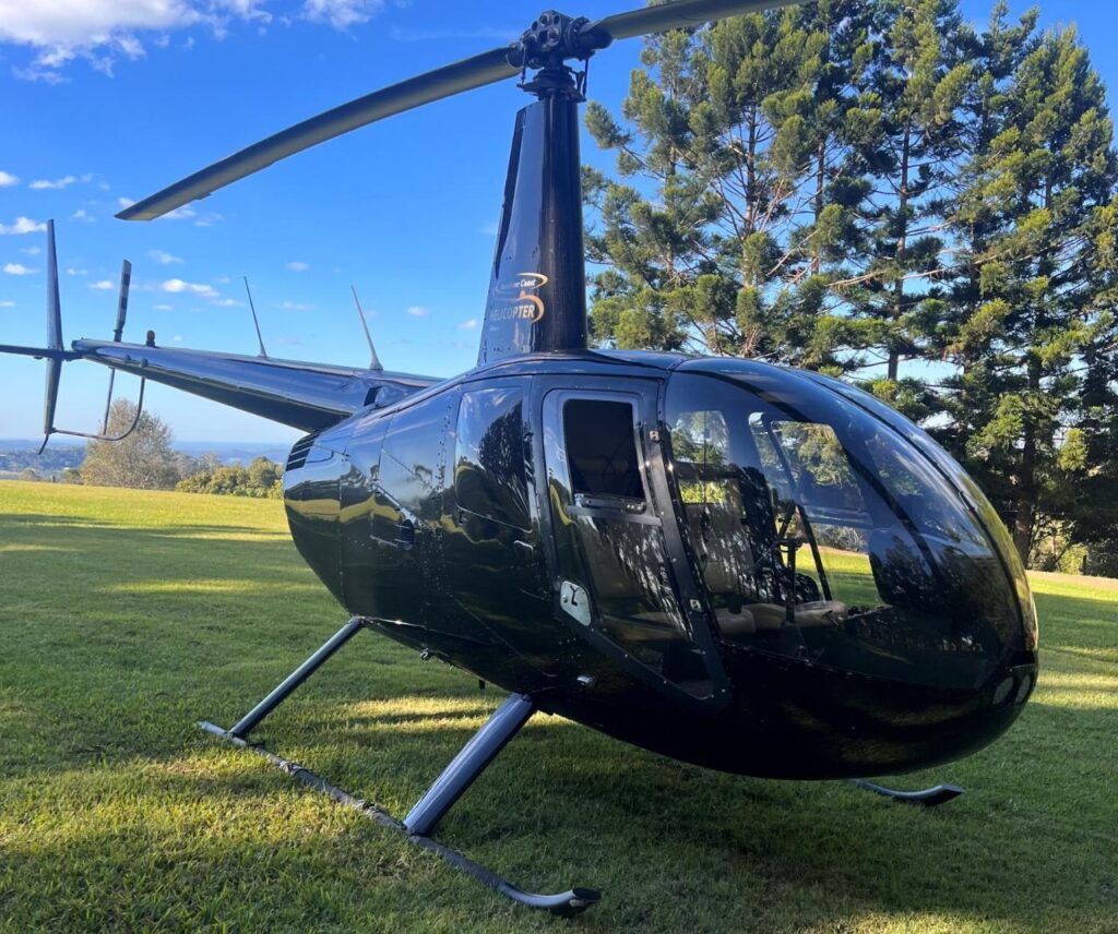 A black helicopter is parked on a grassy field with trees and blue sky in the background.