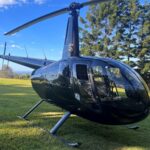A black helicopter is parked on a grassy field with trees and blue sky in the background.