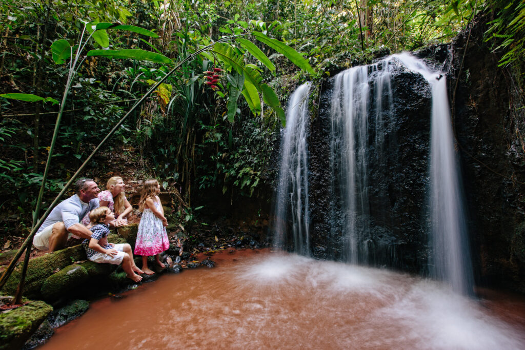 A family of five sits on a rock by a small waterfall in a lush, green forest, watching the water flow into a shallow reddish-brown pool.